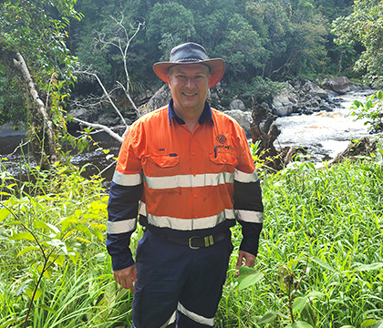 Iain wearing Optravation Energyhigh-visibility orange and navy blue safety workwear and a wide-brimmed hat, standing outdoors in lush green foliage near a river in a forested area.
