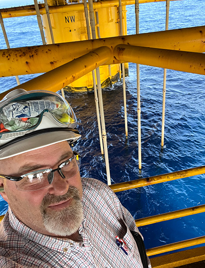 Marty on an offshore platform, wearing safety gear with ocean water and yellow steel structure in the background.