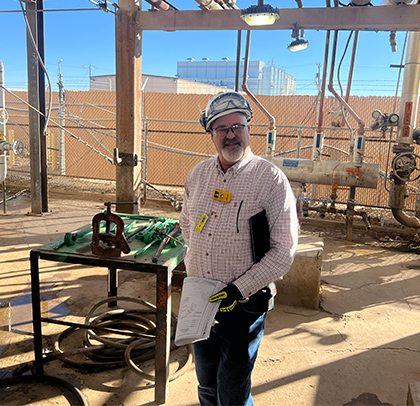 Marty standing in an industrial plant area, wearing safety helmet and holding documents beside equipment and piping.