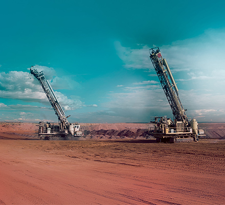 Two large drilling rigs operating in an open‑pit mine, positioned on a wide, dusty bench under a blue sky.